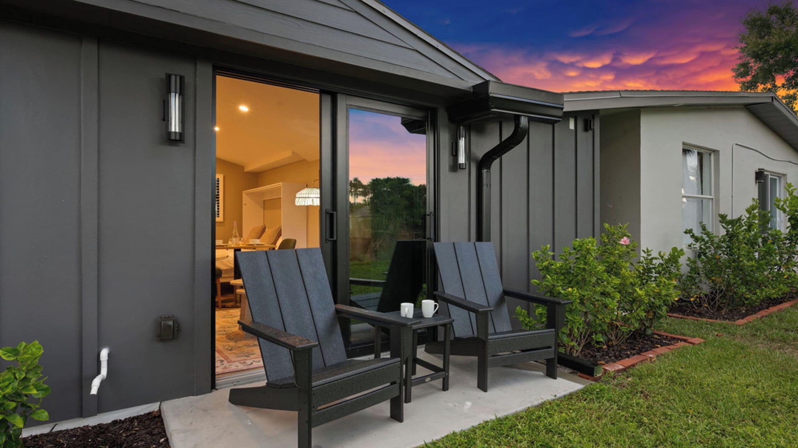 A modern house with a patio area featuring two chairs and a table, a glass door leading to an interior room, and a garden with plants.