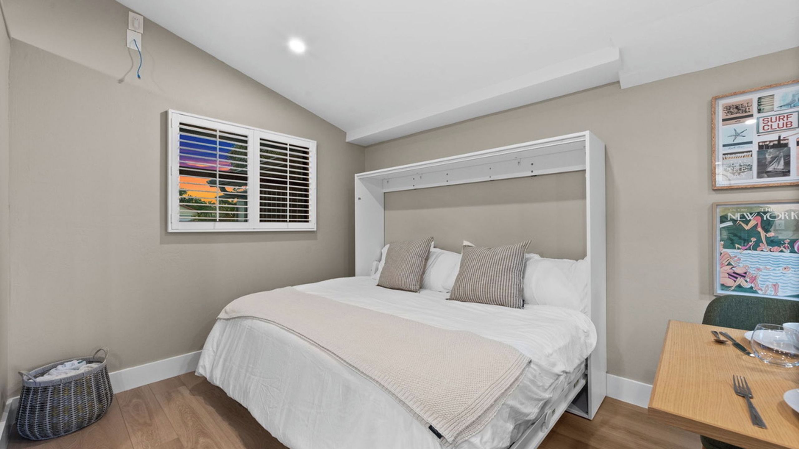 Ein Schlafzimmer mit einem Bett, einem Fenster mit Jalousien, einem an der Wand befestigten Regal und einem kleinen Tisch mit Stuhl.