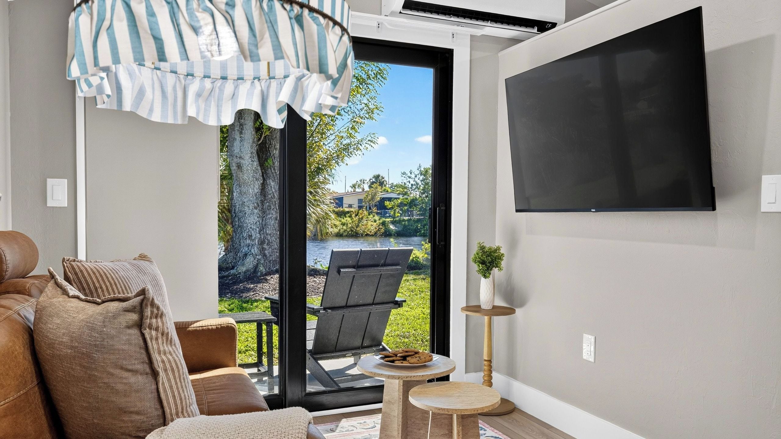 Sala de estar com sofá, mesa de centro, TV fixada na parede e uma porta de vidro deslizante que dá para área externa com cadeira de varanda e mesa pequena.