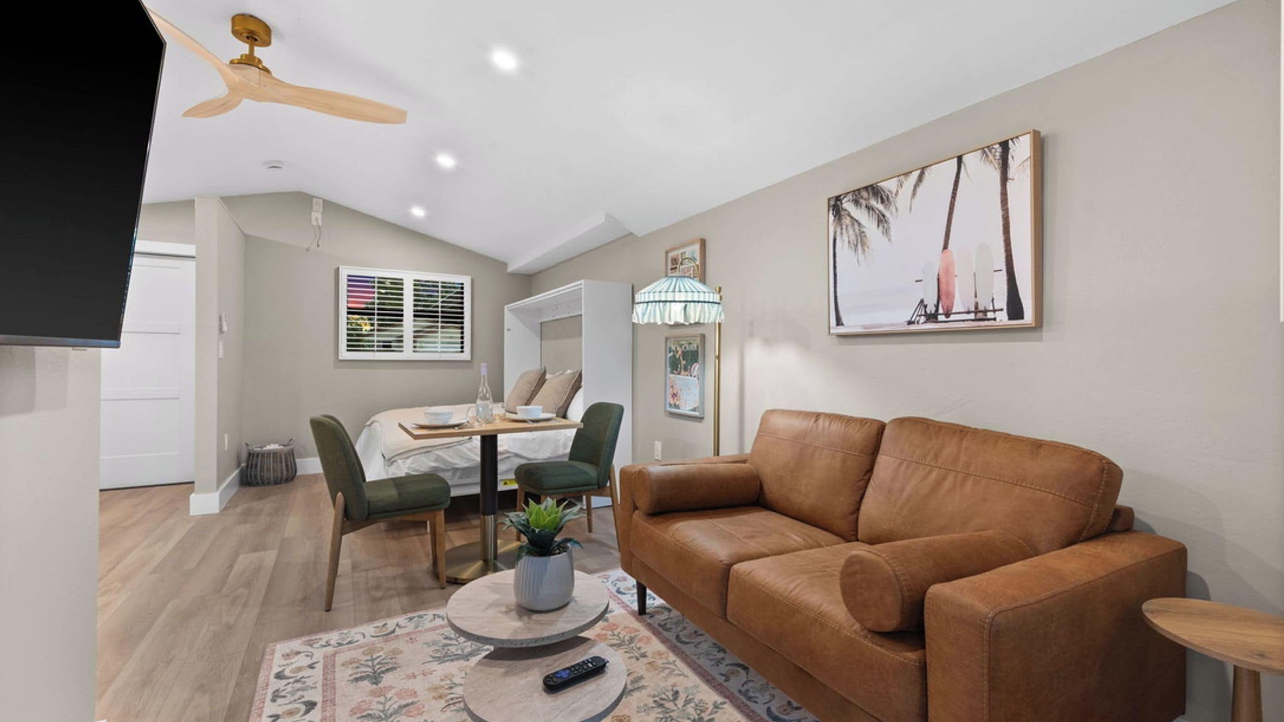 A modern living room with a brown leather sofa, a dining area with a table and chairs, a ceiling fan, and a beach-themed wall art.