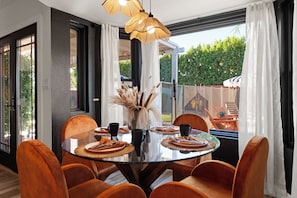 A dining area with a glass table surrounded by chairs, a vase with dried plants, and a view of the outdoors. - Desert Social- Pool, Hot Tub, Game Room & Mini Golf (Scottsdale)