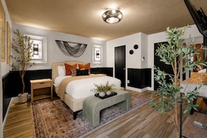 A modern bedroom with a large bed, bedside tables, a nightstand, a decorative wall hanging, a ceiling light fixture, and a potted plant. - Desert Social- Pool, Hot Tub, Game Room & Mini Golf (Scottsdale)
