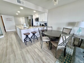 A modern kitchen with white cabinets, a central island with stools, and a dining area with a round table and chairs. - 3BR Gulf Front condo with Amazing Views - sleeps 10 - Pet Friendly (Gulf Shores)