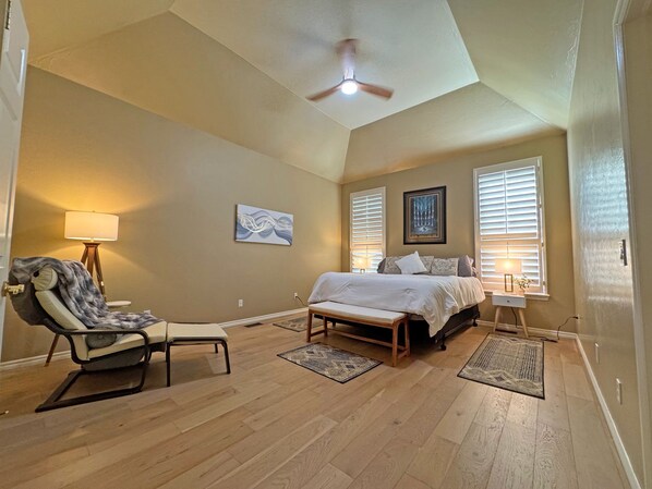 A bedroom with a bed, a chair, a ceiling fan, and two windows with blinds.