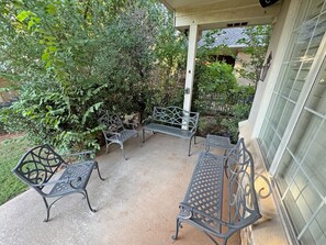 An outdoor patio with metal benches and a concrete floor, surrounded by greenery and a building with a window.