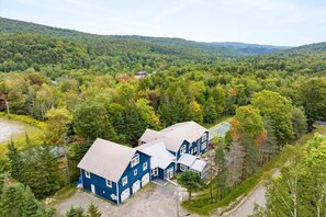 Une grande maison bleue, composée de plusieurs parties, entourée d’une forêt dense et de collines.