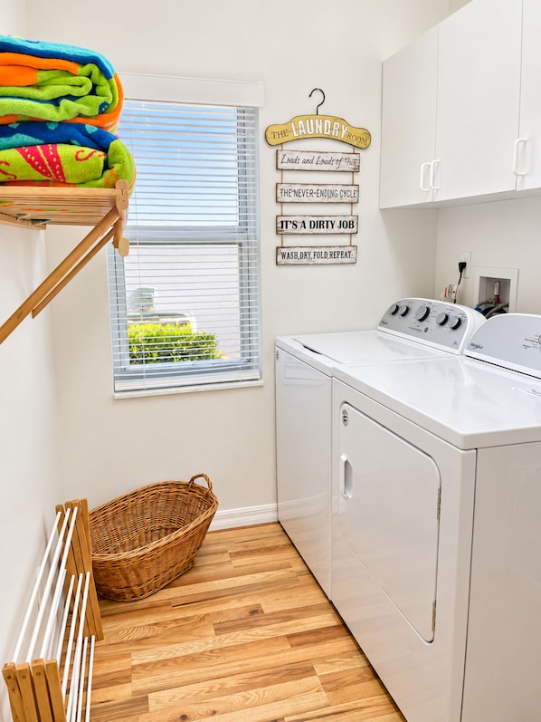 A laundry room with a stacked drying rack, a washing machine, a wicker basket, and a window with blinds.