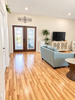 A spacious living room with a wooden floor, a blue sofa, a television on a cabinet, and a decorative wall piece.