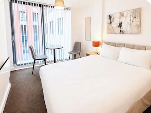 A hotel room with a large bed, a chair, a small table, a lamp, and a view of buildings through the window. - Stylish 2BR | City Centre | Near Northern Quarter (Manchester)