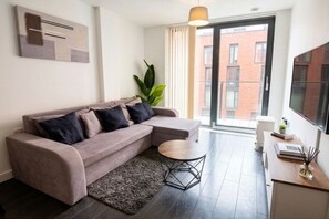 A modern living room with a sectional sofa, a round coffee table, and a view of a brick building through a glass door. - Stylish 2BR | City Centre | Near Northern Quarter (Manchester)