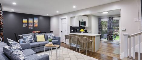A modern living room with a grey sofa, a dining area with a white table, and a kitchen with a microwave.