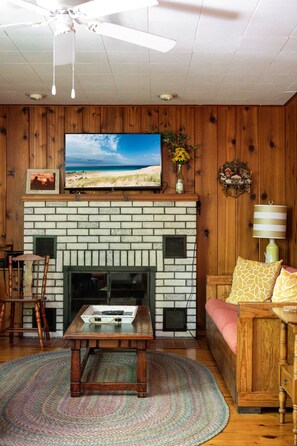 Una sala de estar con chimenea, un televisor montado en la pared, un ventilador de techo y piso de madera.