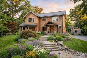 Une maison à deux étages, avec des fondations en pierre, un revêtement extérieur en bois et un jardin aménagé à l’avant, avec un chemin en pierre menant à l’entrée.