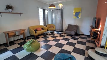 Una sala de estar con piso a cuadros, un sofá, una silla, una mesita, una bolsa verde y un televisor.