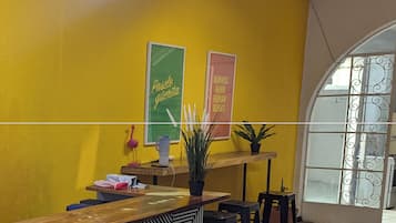 Zona de recepción con mostrador de check-in, un letrero verde y rosa, una planta en maceta y una ventana con rejas decorativas.