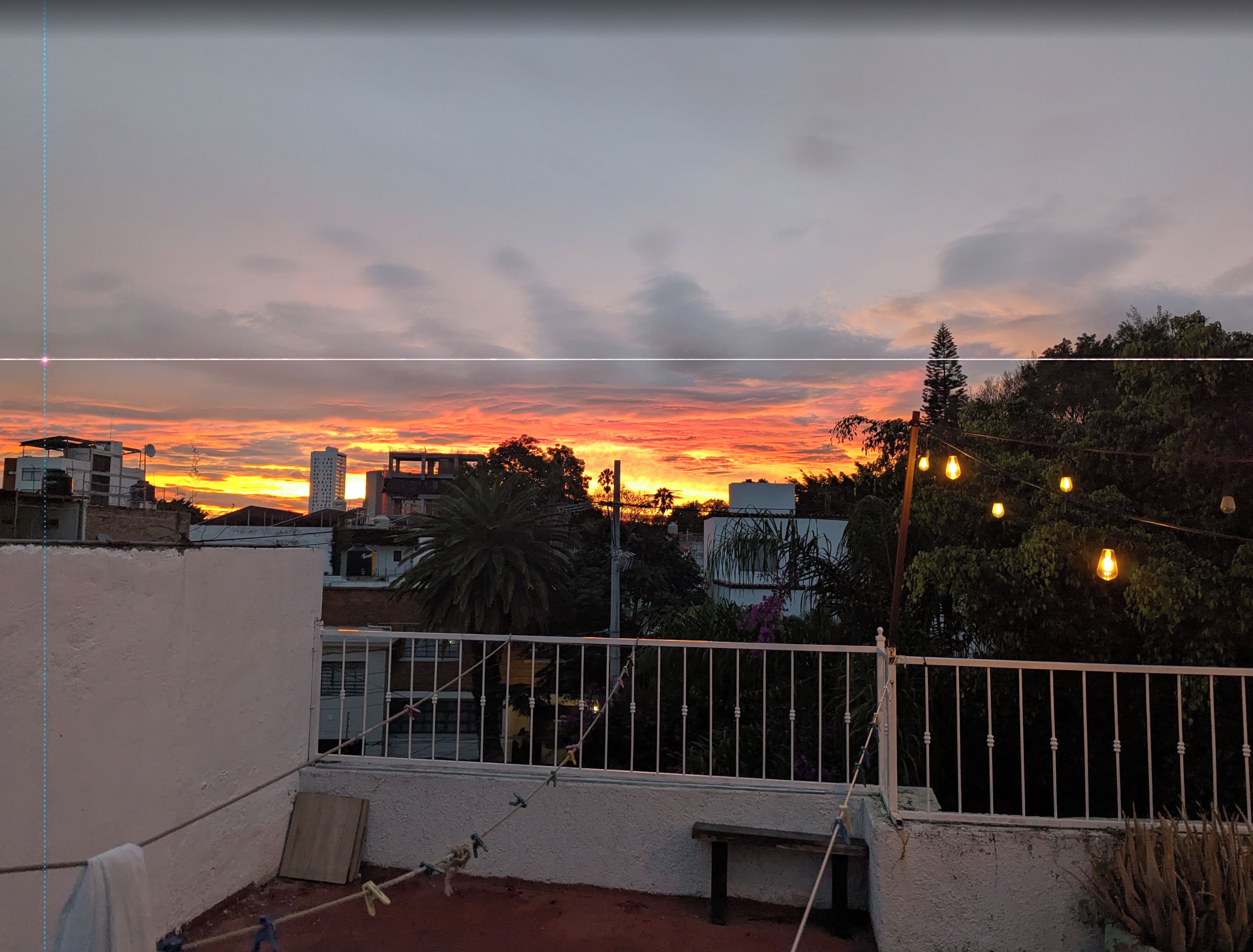 Una azotea con vista a la puesta de sol sobre un paisaje urbano, con edificios y una palmera.