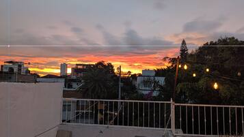Una azotea con vista a la puesta de sol sobre un paisaje urbano, con edificios y una palmera.