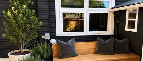A wooden bench with cushions, a potted plant, and a window with a view of a house.