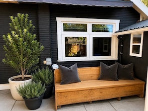A wooden bench with cushions, a potted plant, and a window with a view of a house.