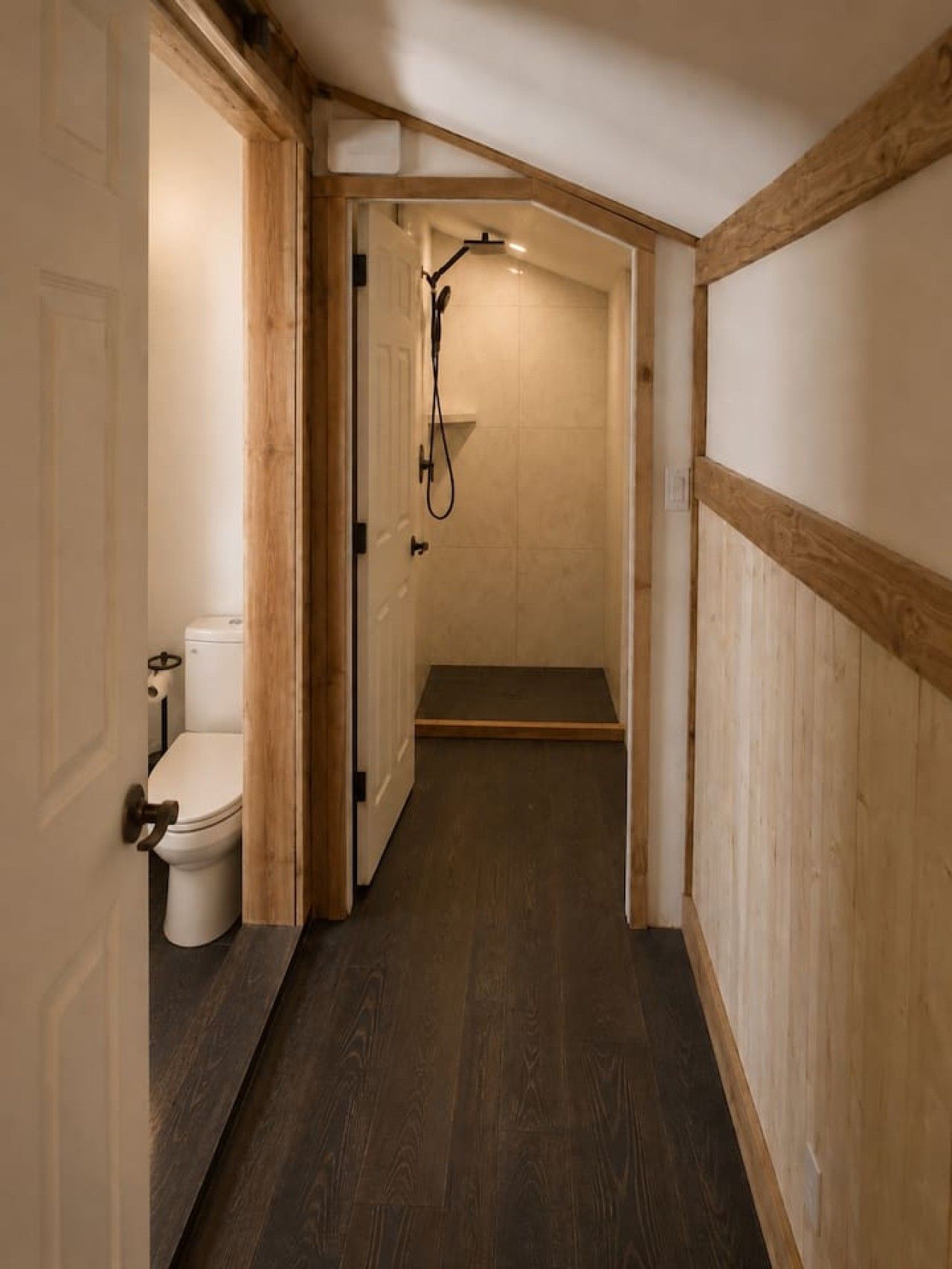 A narrow hallway with a wooden floor leading to a shower area with a showerhead and a toilet.