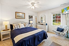 A bedroom with a bed, bedside lamps, a chair, a dresser, a mirror, a ceiling fan, and a TV. - Sandy Blue hot tub, and easy beach access. (Nags Head)