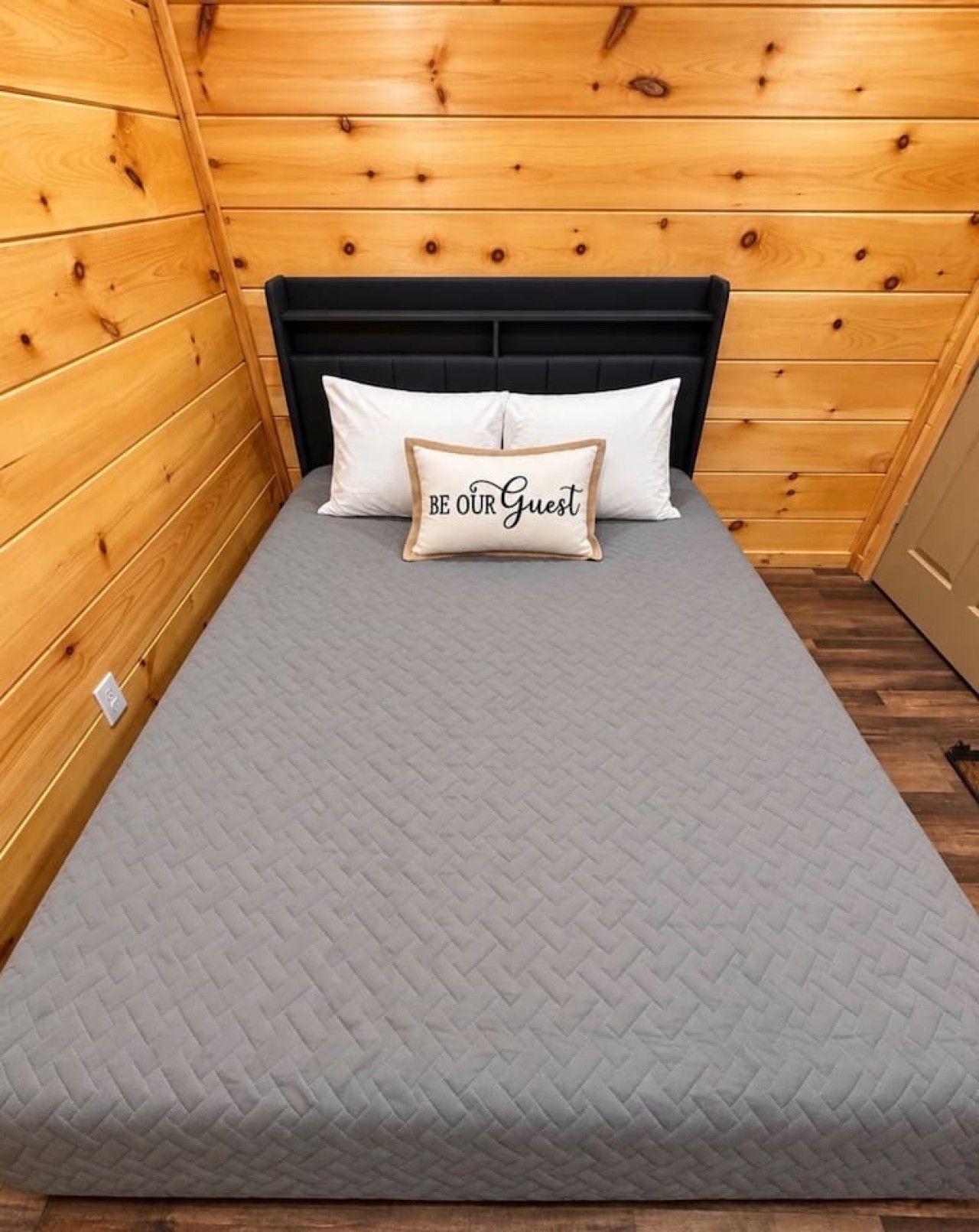 Uma cama de solteiro com colchão cinza acolchoado, cabeceira preta e almofada com a inscrição 'BE OUR GUEST'.