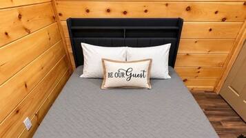 Uma cama de solteiro com colchão cinza acolchoado, cabeceira preta e almofada com a inscrição 'BE OUR GUEST'.