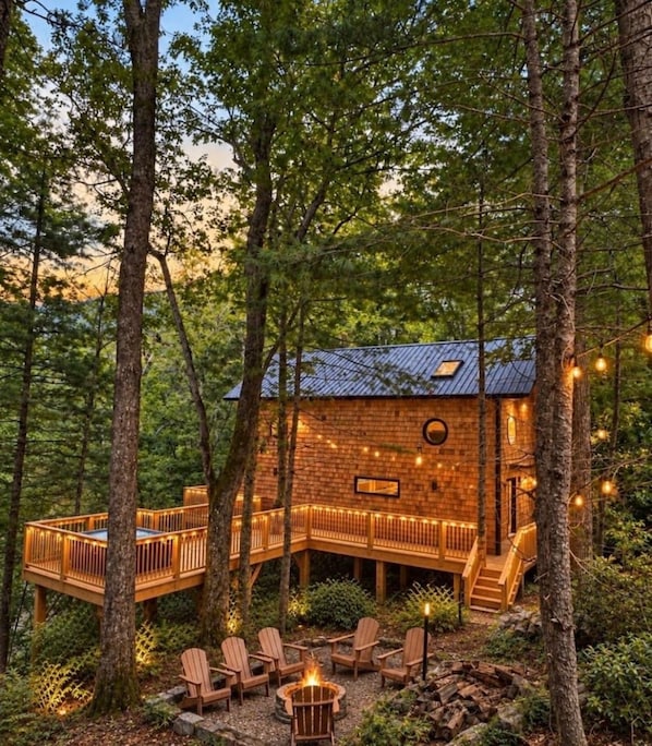 Une cabane dotée d’une terrasse, entourée d’arbres, et aménagée avec un espace salon extérieur et un foyer.