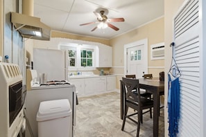 A compact kitchen with white cabinetry, a dining table with chairs, a ceiling fan, and a window with a view of greenery. - Downtown 1BR Home | Walk to Restaurants & Music (Lafayette)