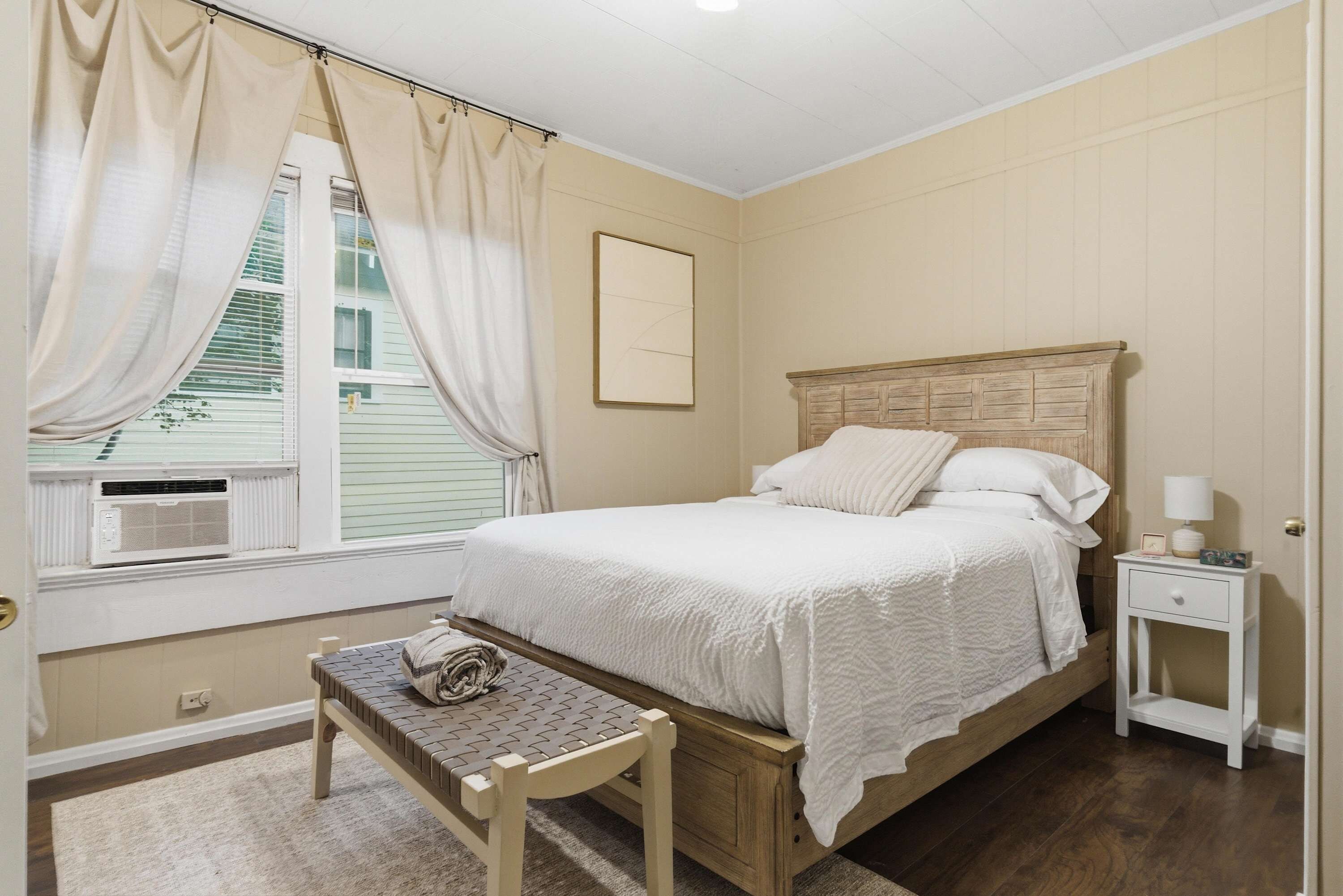 A bedroom with a bed, a bedside table, a nightstand, a window with curtains, and a radiator.