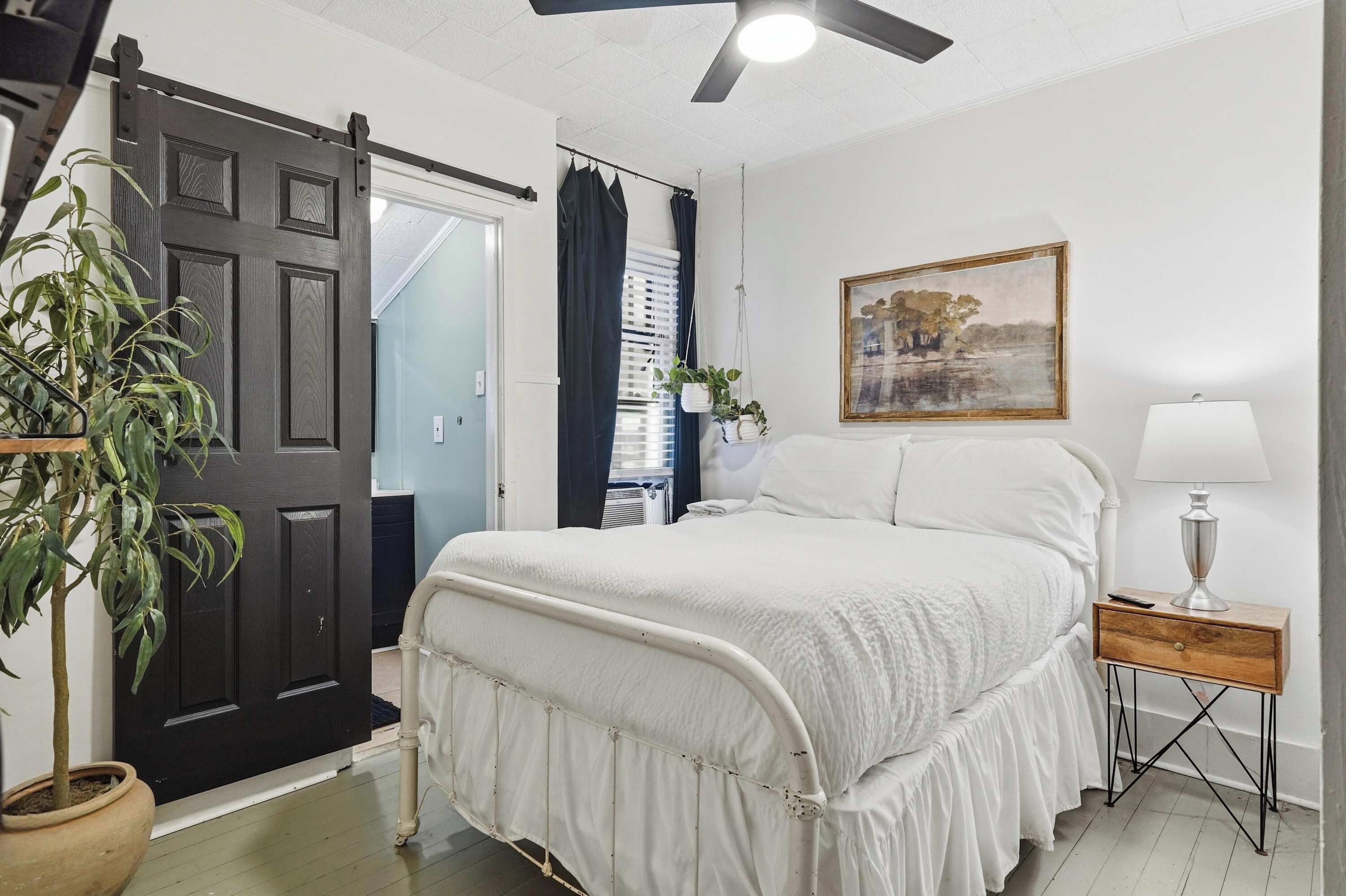 Una camera da letto con un letto, un comodino, una lampada, un quadro, una ventola a soffitto e una porta.