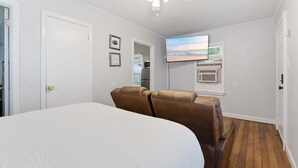 Une chambre à coucher avec un lit, deux fauteuils en cuir marron, un ventilateur de plafond, un téléviseur fixé au mur et une fenêtre avec des stores.