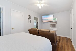 Quarto com uma cama, duas poltronas de couro marrom, um ventilador de teto, uma TV na parede e uma janela com persianas.