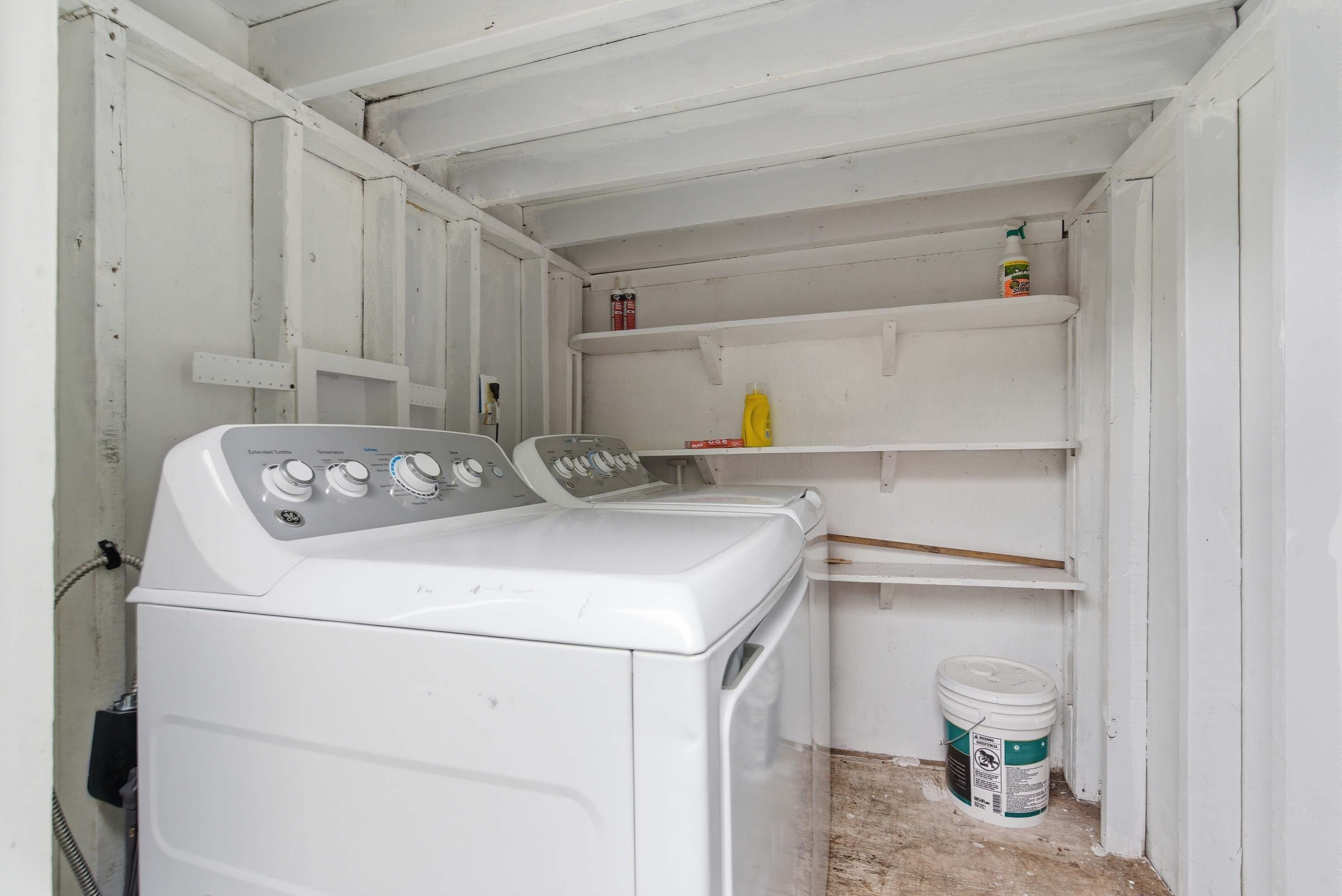 A compact laundry room with a front-loading washing machine, shelves above for storage, and a bucket of detergent on the floor.