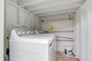 A compact laundry room with a front-loading washing machine, shelves above for storage, and a bucket of detergent on the floor.