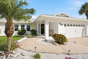 Una casa residencial con garaje, un porche cubierto y un jardín bien cuidado con una palmera y plantas con flores.