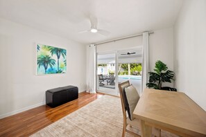Quarto com piso de madeira, um banquinho preto, mesa de jantar de madeira, ventilador de teto e uma janela grande com vista para uma piscina ao ar livre e mobiliário de varanda.