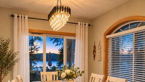 Comedor con mesa dispuesta para seis personas, una araña y vista a un lago al anochecer.
