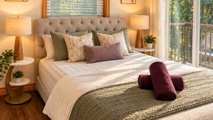 Un dormitorio con cabecera capitonada, cama con múltiples almohadas, mesita de noche con lámpara y vistas a la vegetación por la ventana.