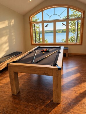 Une table de billard dans une pièce au plancher en bois, avec une grande fenêtre donnant sur un lac.