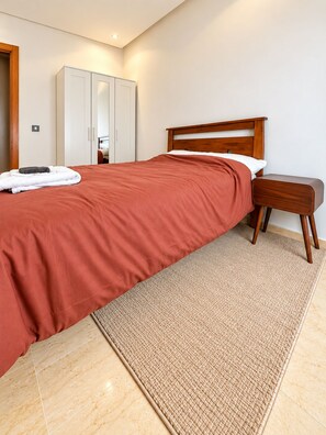 A bedroom with a bed, a wooden nightstand, a white wardrobe, and a bathroom door. - Ideal, 2 bedrooms, 2 bathrooms, fiber optic internet, IPTV, parking, fitness (Dar-el-Beida)