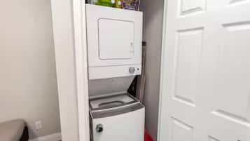A compact laundry area with a washing machine and dryer, a small shelf with detergent, and a red trash bin.
