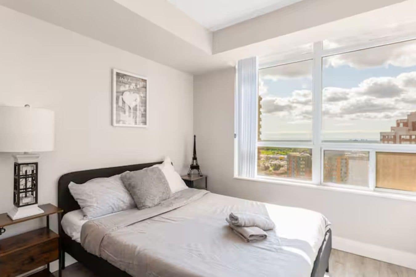 A bedroom with a bed, a nightstand, a lamp, a framed picture, and a window with a view.