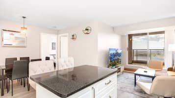A modern living room with a large window, a dining area with a table and chairs, and a kitchen with a granite countertop.