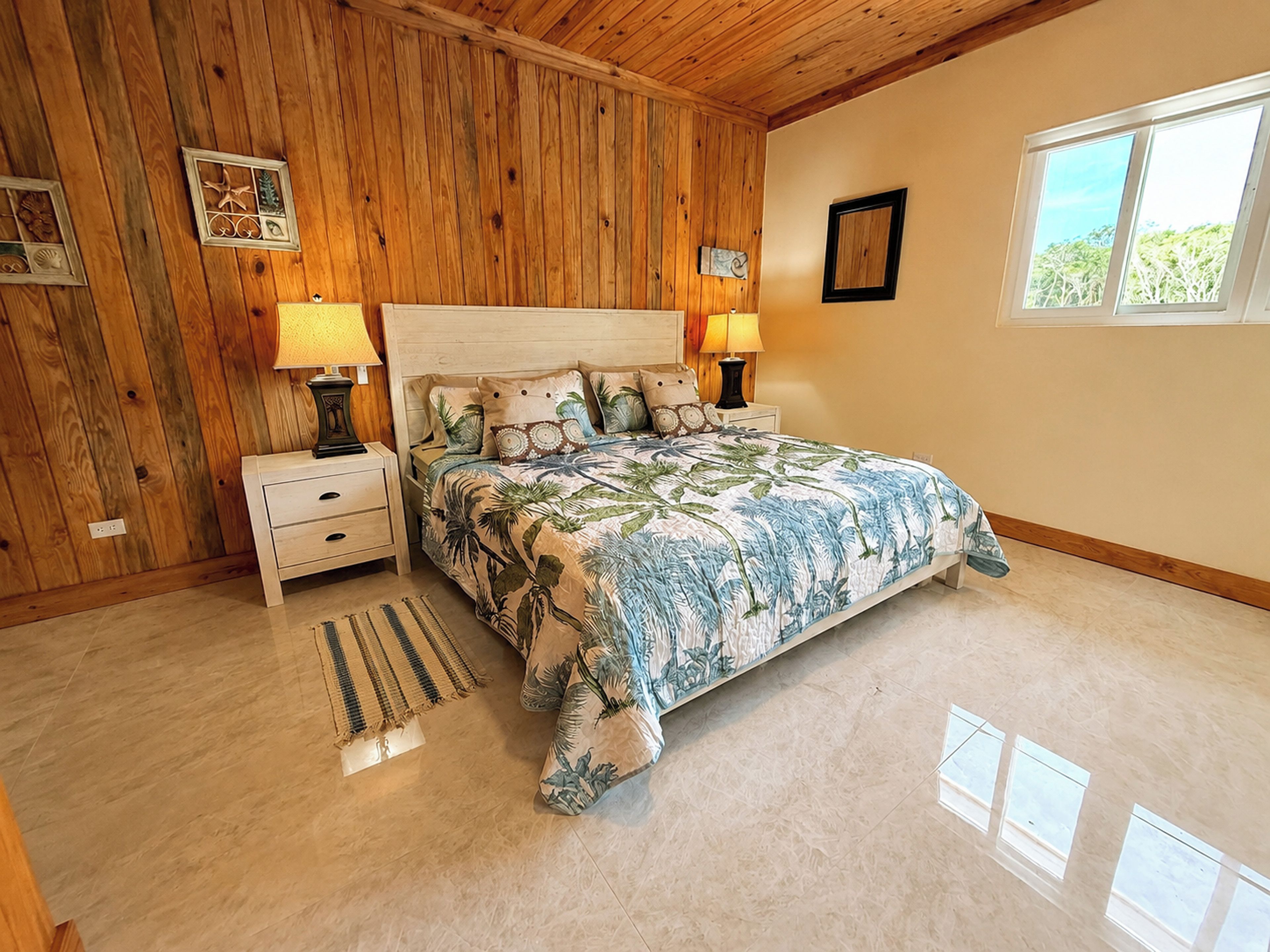 Une chambre avec un plafond en bois, un lit recouvert d’une courtepointe à motifs floraux, deux tables de chevet avec des lampes, une fenêtre donnant sur un beau paysage et un petit tapis sur le sol.