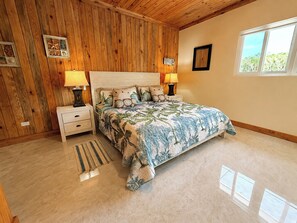 Een slaapkamer met een houten plafond, een bed met een bloemenprint, twee nachtkastjes met lampen, een raam met uitzicht, en een klein vloerkleed.