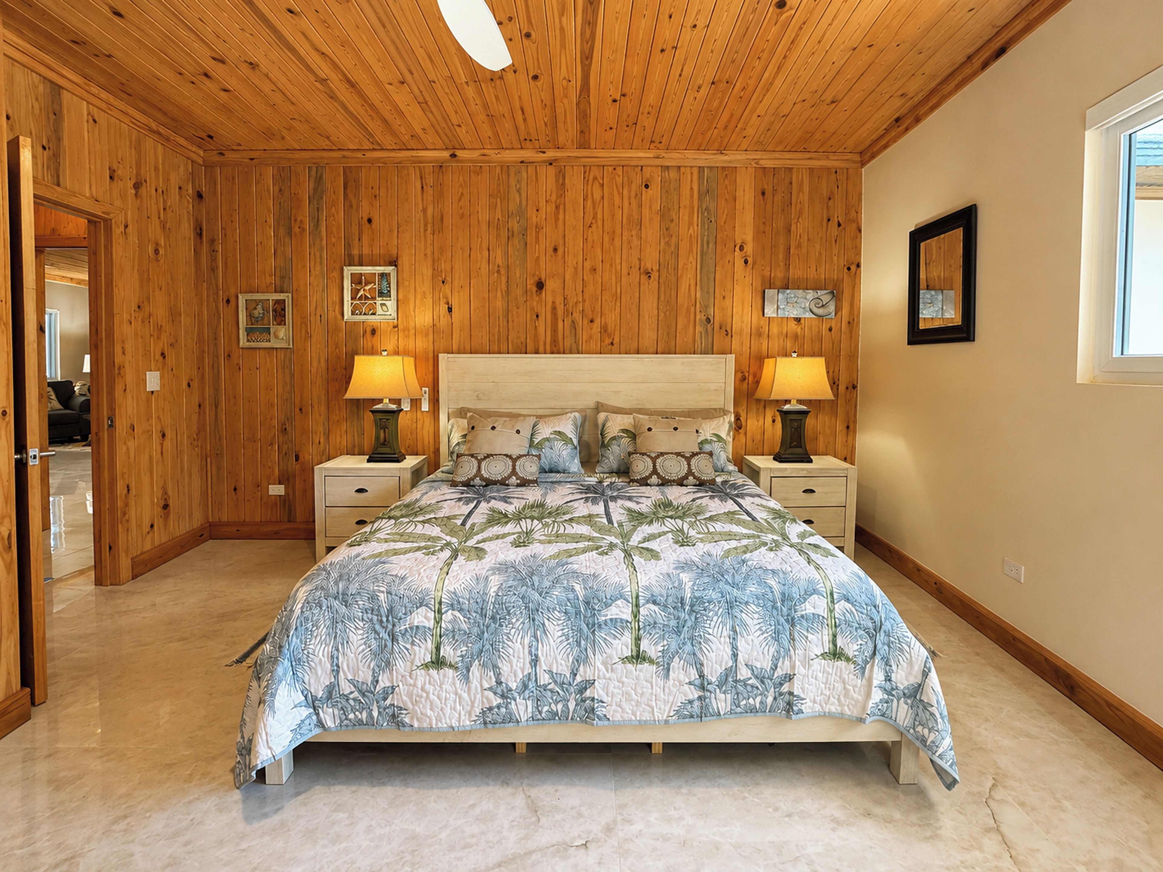 Un dormitorio con paredes de madera, una cama grande con colcha floral, dos mesitas de noche con lámparas y cuadros enmarcados en las paredes.