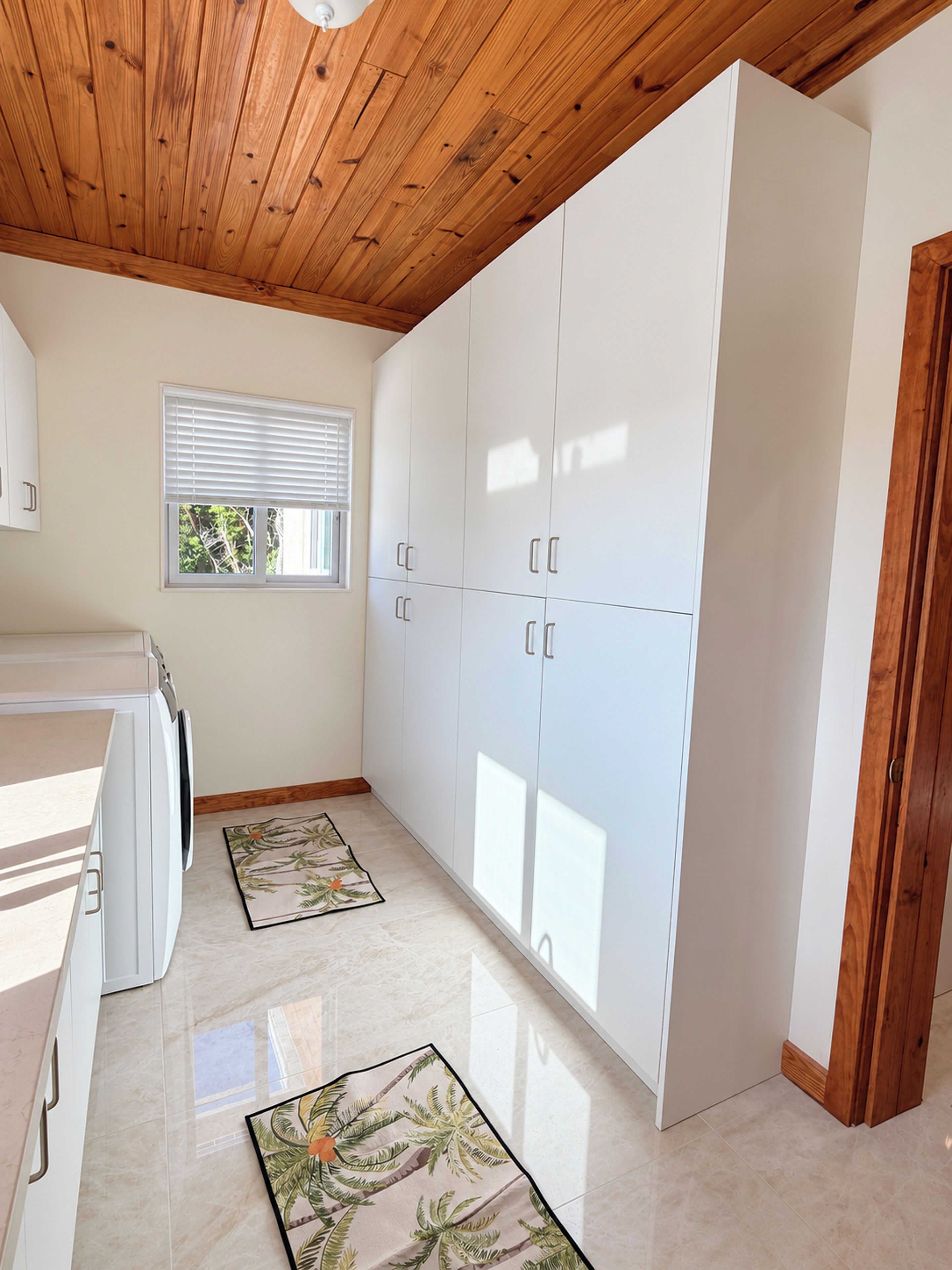 Una cocina con armarios blancos, techo de madera, ventana con persianas y alfombra estampada.