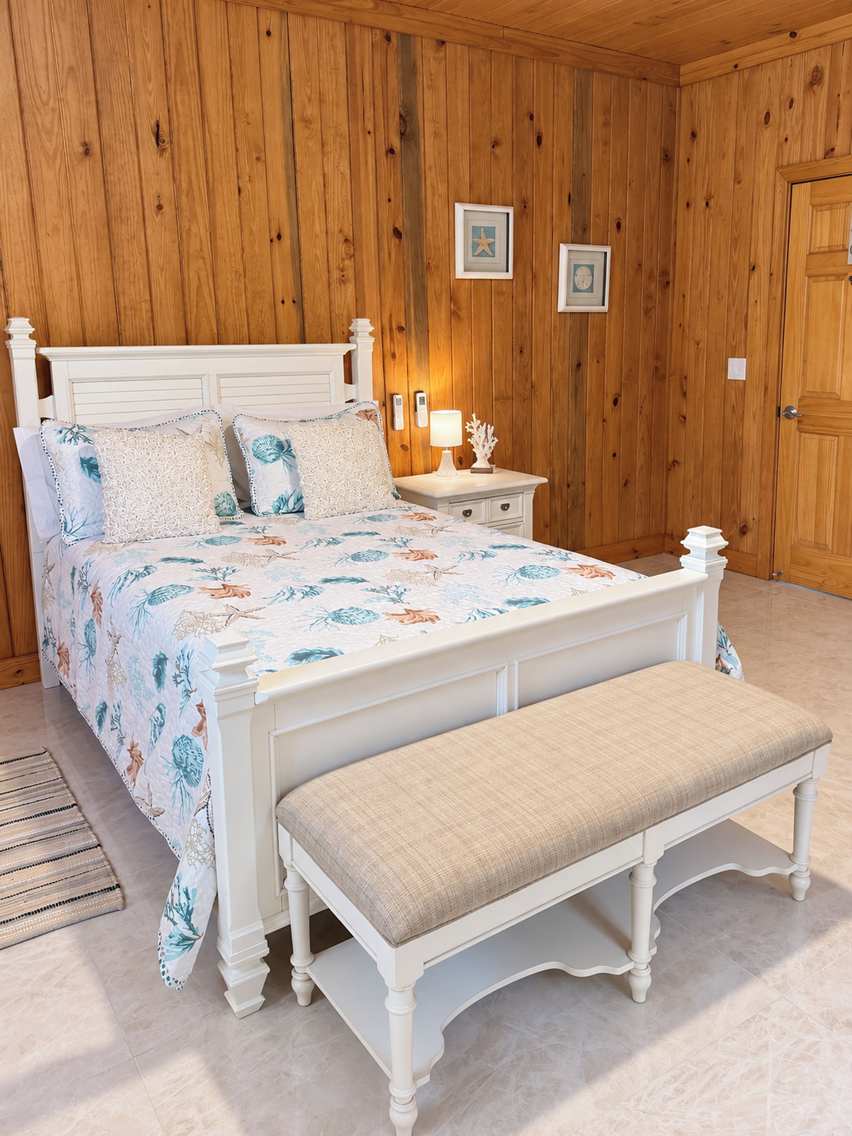 A bedroom with a wooden paneled wall, a white bed with floral bedding, a bedside table with a lamp, and a bench.