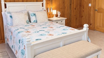 A bedroom with a wooden paneled wall, a white bed with floral bedding, a bedside table with a lamp, and a bench.
